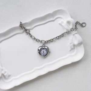May include: A silver heart-shaped watch with a chain bracelet. The watch is on a white ceramic tray.