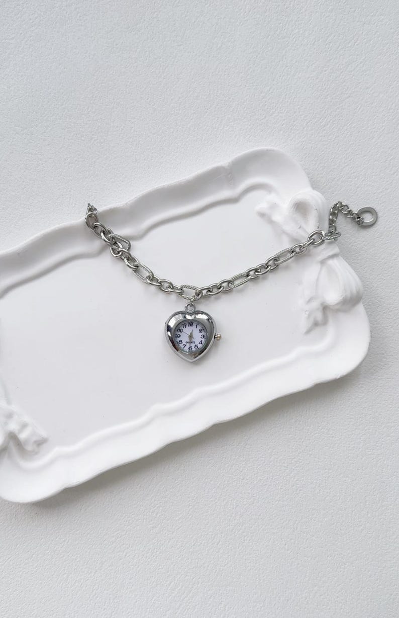May include: A silver heart-shaped watch with a chain bracelet. The watch is on a white ceramic tray.