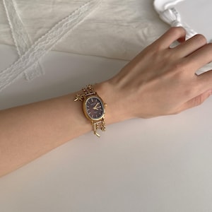 May include: Gold-toned wristwatch with an oval face and a dark brown dial. The watch features delicate charms and is worn on a wrist. The watch has a gold band and a star charm. The watch is a fashion accessory.