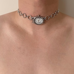 May include: Silver-toned watch choker necklace. The watch face has a white background with black Roman numerals and hands. The necklace is composed of small, connected rings.