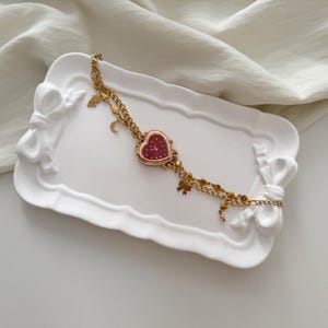 May include: A gold-toned watch with a heart-shaped face and a burgundy dial rests on a white, bow-shaped tray. The watch features a double chain with small charms, including a crescent moon and a bow.