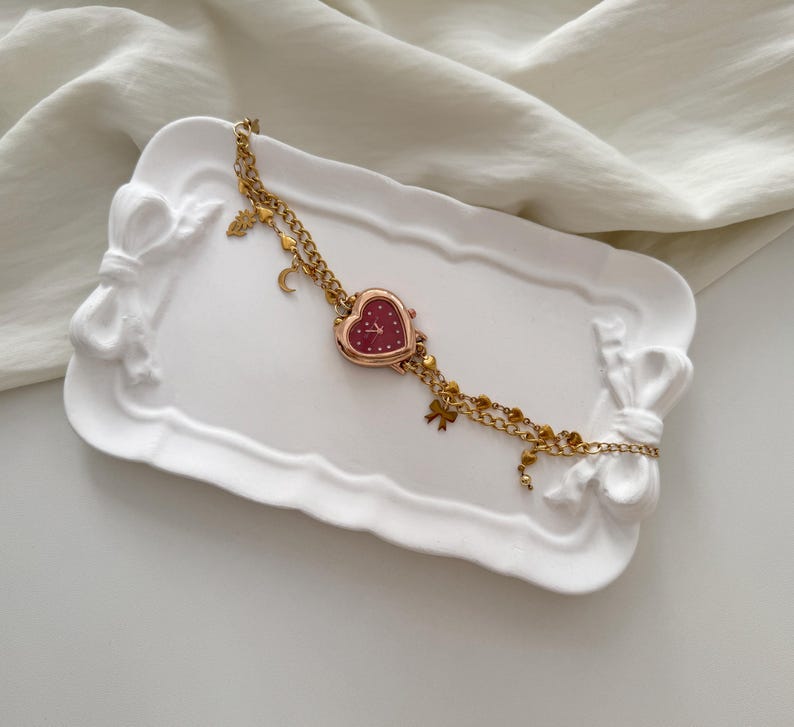 May include: A gold-toned watch with a heart-shaped face and a burgundy dial rests on a white, bow-shaped tray. The watch features a double chain with small charms, including a crescent moon and a bow.
