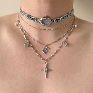 Pode incluir: Um conjunto de colares de prata em camadas. A camada superior é um choker de relógio com mostrador azul claro. Abaixo, duas correntes delicadas com pingentes de estrela, lua e cruz. As joias são exibidas em um fundo neutro.