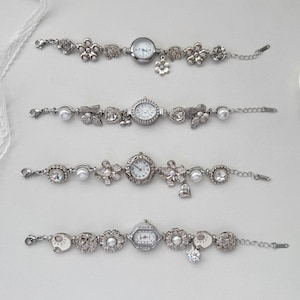 May include: Four silver bracelet watches with floral and gem embellishments. Each watch has a small, round or hexagonal face with a white dial and black numerals. The bracelets incorporate pearls and dangling charms.