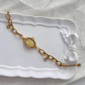 May include: A gold-coloured watch with an oval face and a chain-link bracelet. The watch face displays gold numerals and hands. The bracelet features a series of large, textured links and small, dangling charms. The watch is displayed on a white, decorative tray.