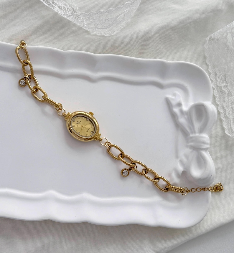 May include: A gold-coloured watch with an oval face and a chain-link bracelet. The watch face displays gold numerals and hands. The bracelet features a series of large, textured links and small, dangling charms. The watch is displayed on a white, decorative tray.