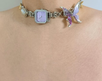 Könnte beinhalten: Eine silberne Kettenkette mit einem lila Schmetterlingsanhänger, einer silbernen Uhr mit einem lila Zifferblatt und einer Perle. Die Kette wird am Hals einer Person getragen.
