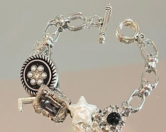 Op de afbeelding: Een zilveren armband met verschillende bedels, waaronder een zwarte knoop met witte parels, een helder kristal, een witte ster en een zwarte steen met strass.
