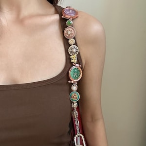 Puede incluir: Una correa marrón con múltiples abalorios decorativos y esferas de reloj. La correa está unida a un bolso de mano rojo.