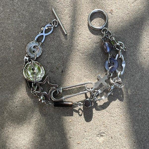May include: A silver charm bracelet featuring a range of charms, including buttons, stars, a safety pin, and a pearl. The bracelet has a toggle clasp.