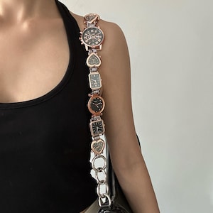May include: A rose gold chain bag strap with multiple small watch faces attached. The strap is made of metal and has a heart-shaped design. The bag strap is attached to a black handbag.