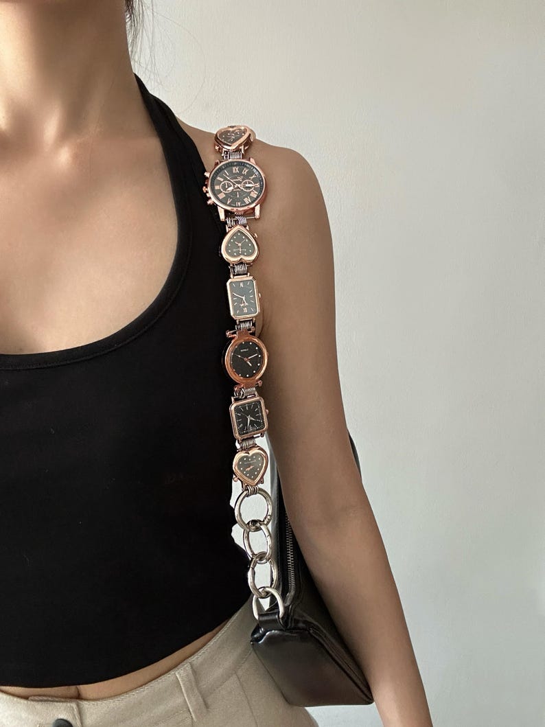 May include: A rose gold chain bag strap with multiple small watch faces attached. The strap is made of metal and has a heart-shaped design. The bag strap is attached to a black handbag.
