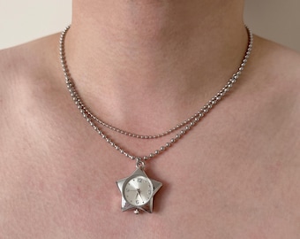 Op de afbeelding: Een zilverkleurige ketting met een stervormige hanger die als horloge fungeert. De wijzerplaat van het horloge heeft zwarte cijfers en wijzers. De ketting is gemaakt van kleine, ronde kralen.
