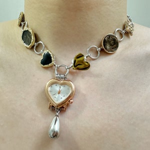 Peut inclure: Un collier en argent et or avec un pendentif de montre en forme de cœur. Le collier présente plusieurs breloques en forme de cœur, y compris des pierres noires, dorées et transparentes.