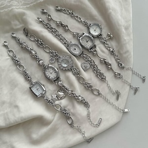 May include: Six silver wristwatches with diverse chain styles and watch faces. The watches have different shapes, including round, oval, and square. Some feature heart-shaped charms and decorative links. The watches are displayed on a white fabric.