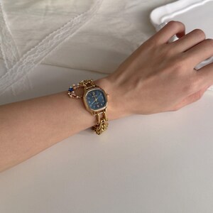 May include: A gold-toned wristwatch with a rectangular face and a dark blue dial. The watch has a gold chain-link band and is adorned with a small charm. The watch is on a wrist.