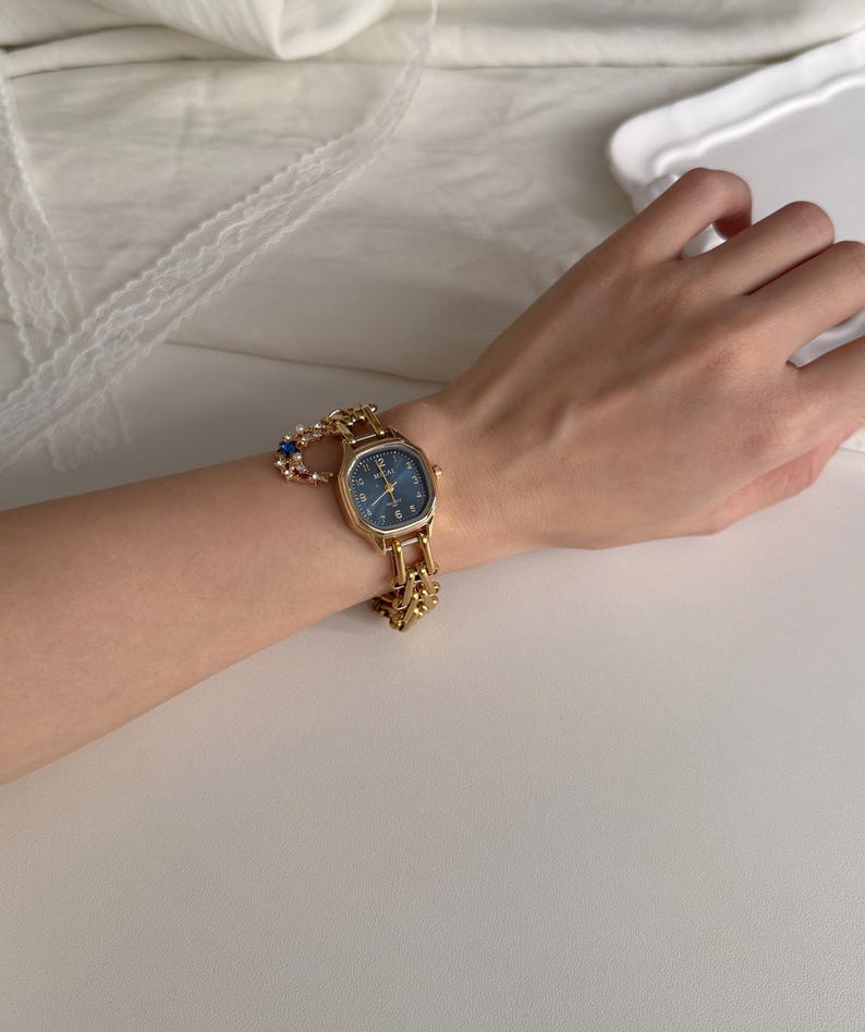 May include: A gold-toned wristwatch with a rectangular face and a dark blue dial. The watch has a gold chain-link band and is adorned with a small charm. The watch is on a wrist.