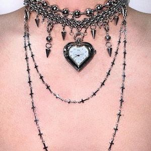 Puede incluir: Un collar de plata con un dije en forma de corazón que contiene una esfera de reloj. El dije está unido a un collar de cadena de capas con un collar gargantilla de plata con púas.