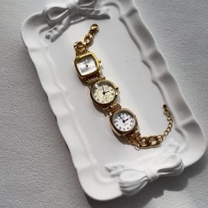 May include: A gold-tone watch bracelet with four different watch faces. The bracelet is displayed on a white ceramic tray with a bow design.