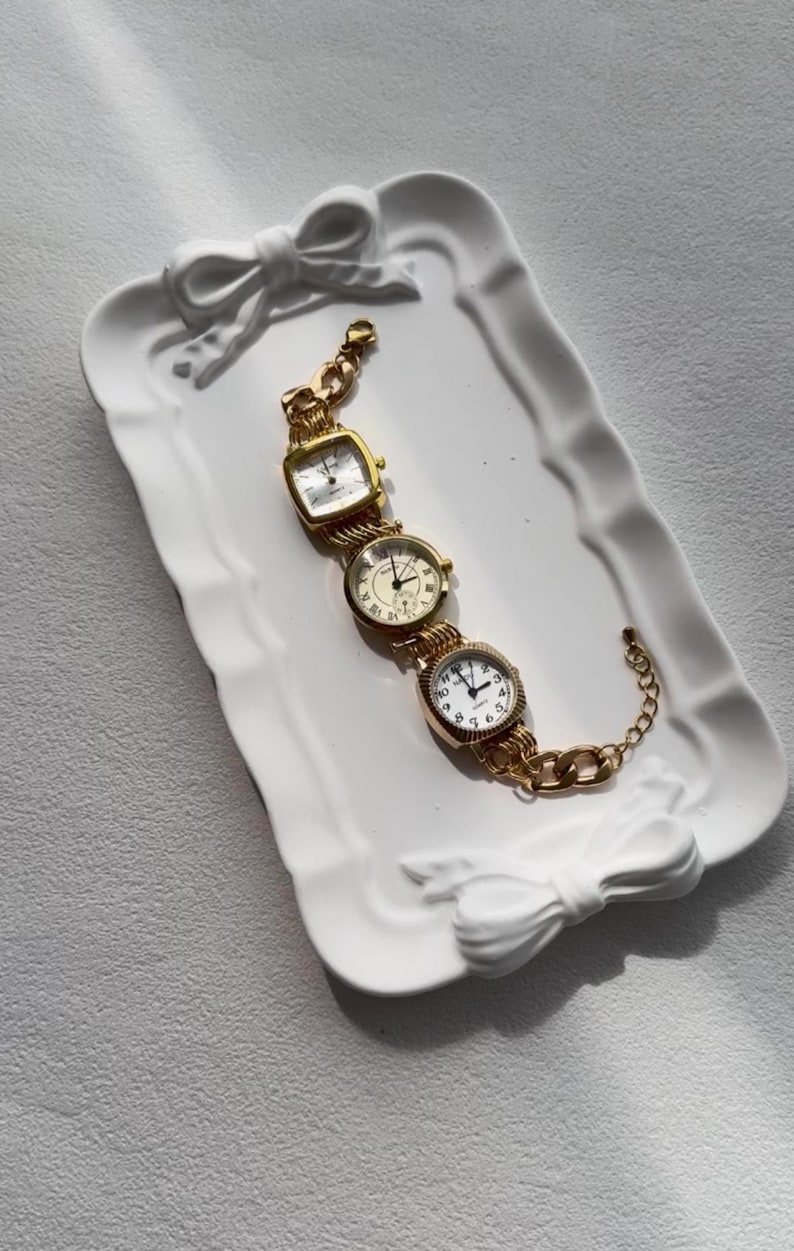 May include: A gold-tone watch bracelet with four different watch faces. The bracelet is displayed on a white ceramic tray with a bow design.