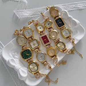 Puede incluir: Una colección de relojes dorados de estilo vintage con varias formas de esfera, incluyendo redondas, cuadradas y rectangulares. Las esferas de los relojes presentan diferentes colores, como blanco, verde y negro. Los relojes se exhiben en una bandeja decorativa blanca.