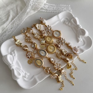 May include: Several gold-toned, decorative wristwatches with various embellishments, including floral, heart, and gemstone accents. The watches have small, round or oval faces. They are displayed on a white, bow-shaped tray.
