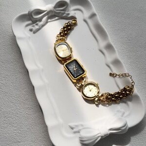 May include: A gold-tone watch with three square faces on a gold chain bracelet. The watch is on a white ceramic tray with a bow design.