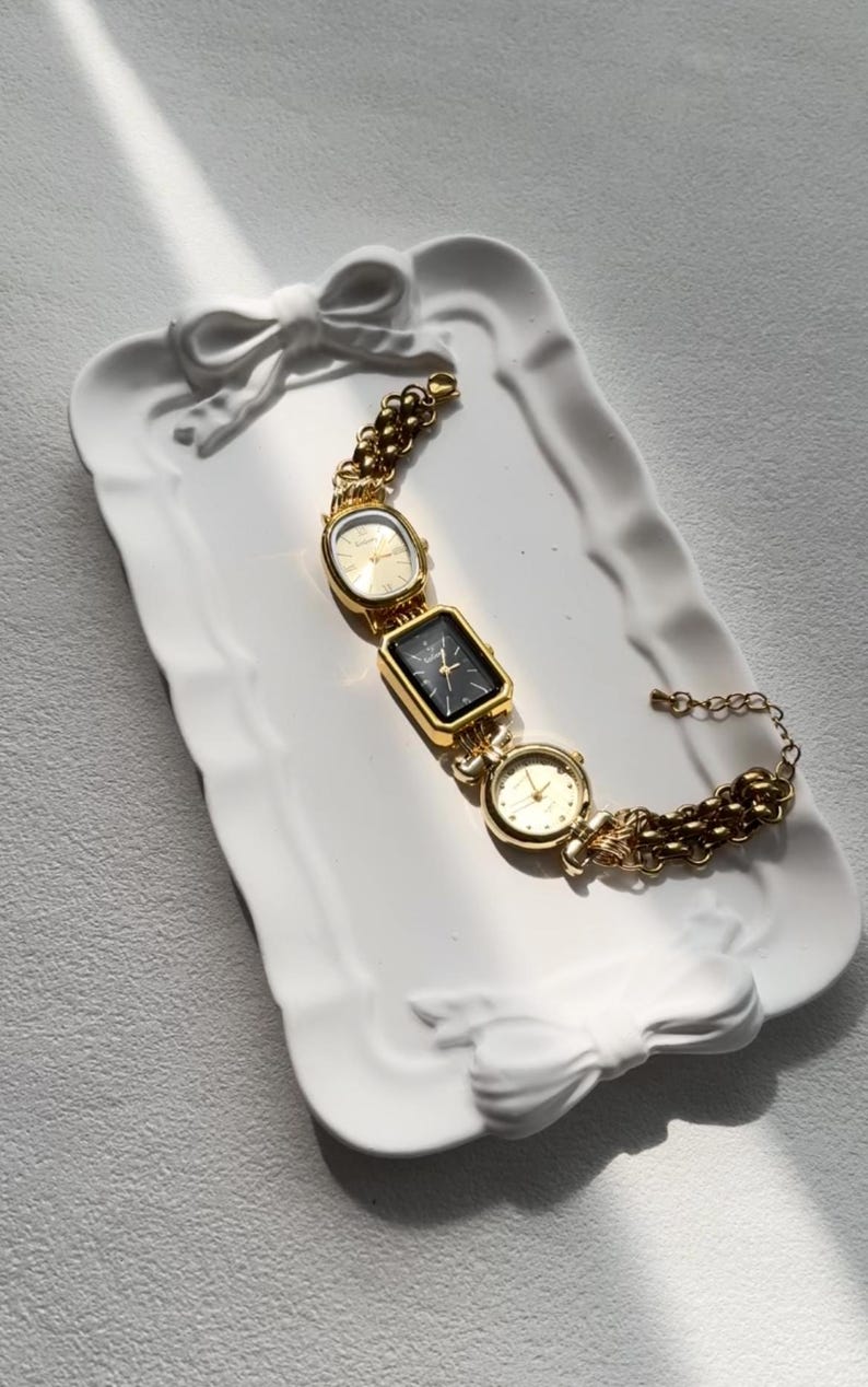May include: A gold-tone watch with three square faces on a gold chain bracelet. The watch is on a white ceramic tray with a bow design.