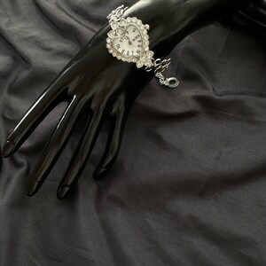 May include: Silver heart-shaped watch with a white face and rhinestone accents. The watch has a chain link bracelet.