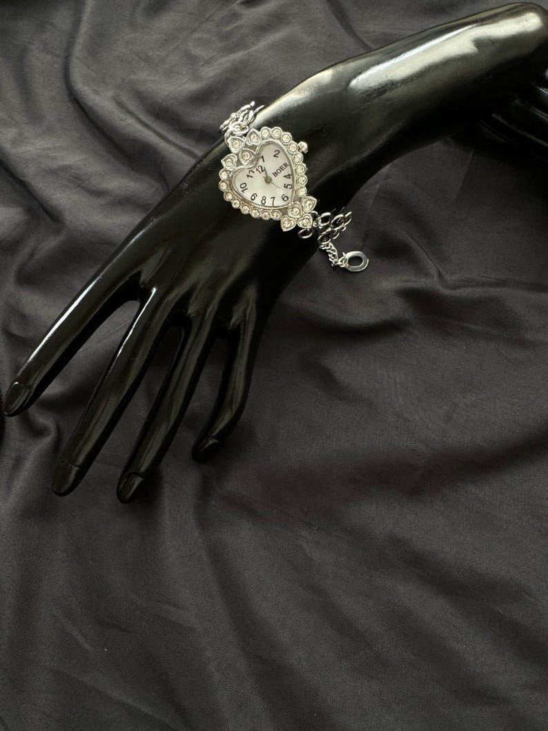 May include: Silver heart-shaped watch with a white face and rhinestone accents. The watch has a chain link bracelet.