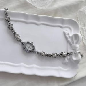 May include: A silver bracelet watch with an oval face and a white dial. The watch band has a delicate design with heart-shaped links and small, round embellishments. The watch is displayed on a white, decorative tray.