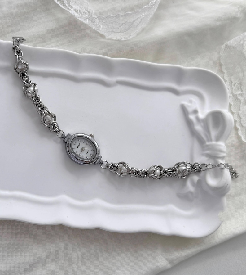 May include: A silver bracelet watch with an oval face and a white dial. The watch band has a delicate design with heart-shaped links and small, round embellishments. The watch is displayed on a white, decorative tray.
