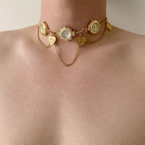 Puede incluir: Un collar gargantilla dorado con múltiples esferas de reloj pequeñas y dijes en forma de corazón. El collar tiene un diseño de cadena delicado con elementos colgantes. Las esferas del reloj tienen esferas de color claro.