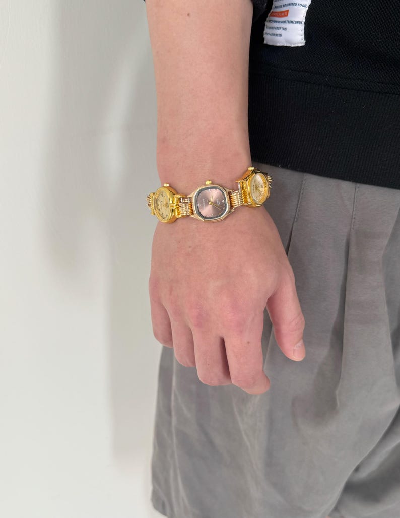 May include: A gold-toned watch bracelet with multiple watch faces. The watch faces are all different sizes and shapes. The bracelet is worn on a person's wrist.