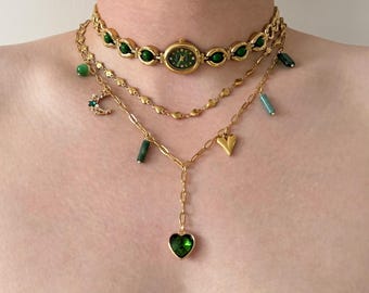 May include: A layered gold necklace set featuring a watch choker with green accents, a delicate chain with a crescent moon charm, and a pendant necklace with a green heart-shaped stone. The necklaces have various green and gold charms.