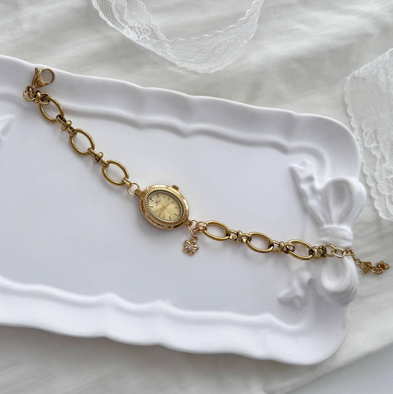 May include: A gold wristwatch with an oval face and a chain-link bracelet. The watch has a small charm and is displayed on a white decorative tray. The watch face is a light gold colour.