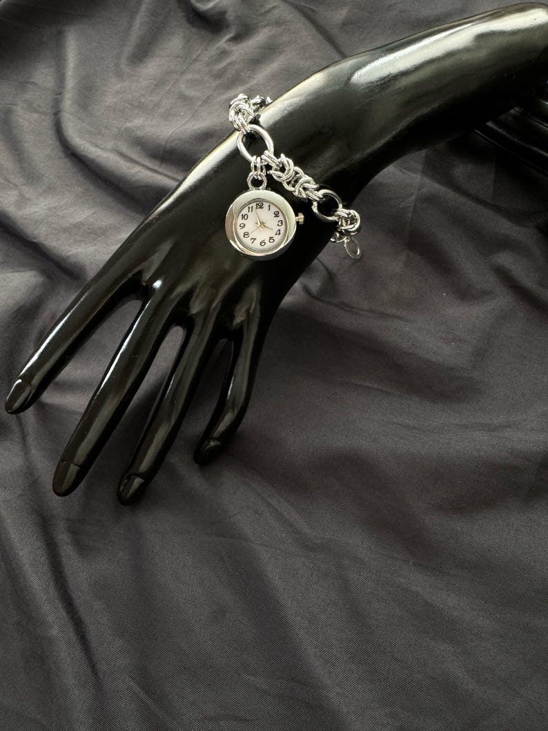 May include: A silver watch with a white face and black numbers on a silver chain bracelet.