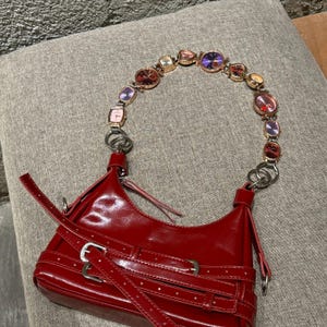 May include: A red leather handbag with a unique watch strap featuring colorful jeweled accents. The bag has a flap closure and multiple straps with buckles.