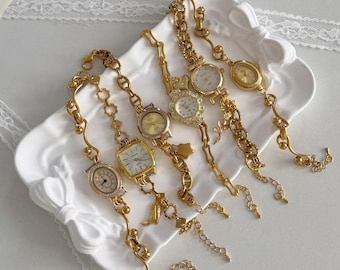 Könnte beinhalten: Sechs goldfarbene Armbanduhren im Vintage-Stil mit verschiedenen Zifferblattformen und Ketten-Designs werden auf einer weißen, bogenförmigen Schale präsentiert. Die Uhren haben runde, ovale und quadratische Zifferblätter mit filigranen Details und Verzierungen.