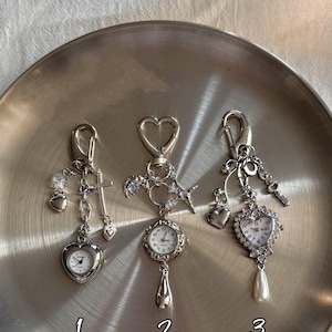Könnte beinhalten: Drei silberfarbene Metallschlüsselanhänger mit Zifferblättern und Anhängern. Schlüsselanhänger 1 hat eine herzförmige Uhr, ein Kreuz und ein Herz. Schlüsselanhänger 2 hat eine runde Uhr, einen Mond und einen Stern. Nummer 3 hat eine herzförmige Uhr mit Kristallen und einer Perle.