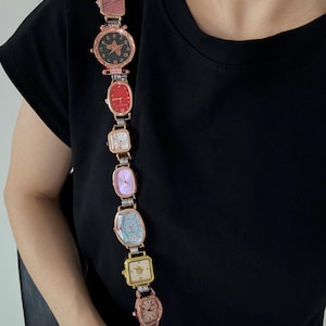 May include: A decorative strap composed of multiple vintage-style wristwatches in various shapes and colors, including rose gold, red, purple, and blue, connected by silver-toned metal links, featuring a circular ring at the bottom end.