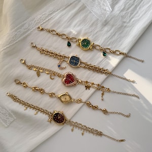 May include: Five gold-toned wristwatches with various colored watch faces, including green, blue, red, and gold. Each watch features a unique design, with charms and decorative chains. The watches are displayed on a white surface.
