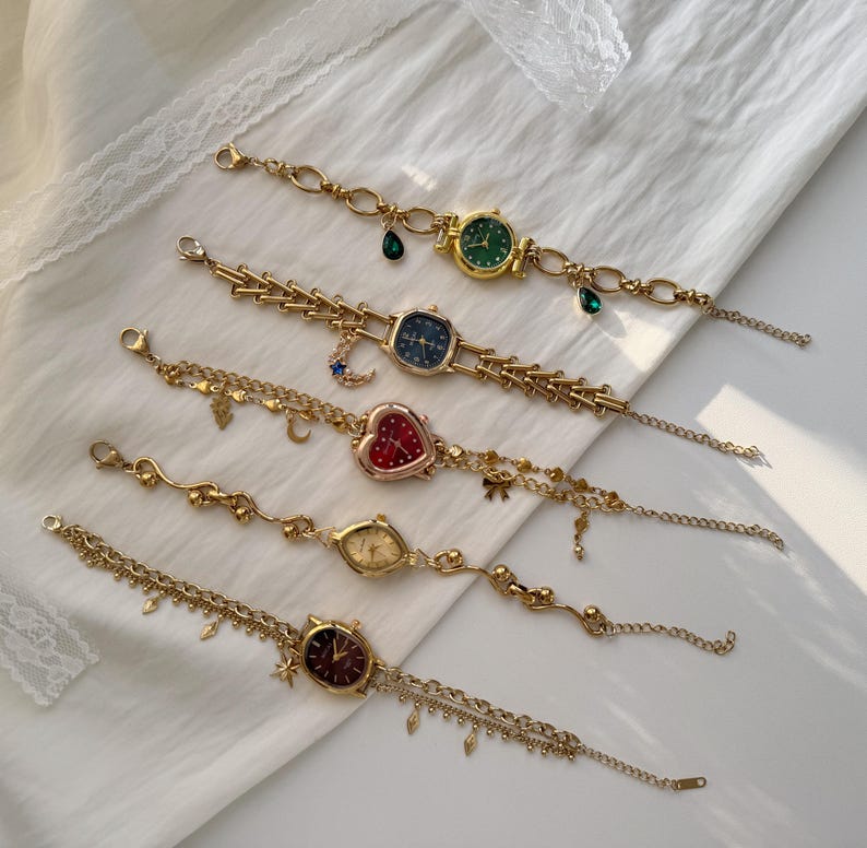 May include: Five gold-toned wristwatches with various colored watch faces, including green, blue, red, and gold. Each watch features a unique design, with charms and decorative chains. The watches are displayed on a white surface.