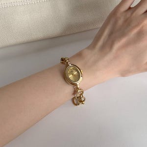 May include: A gold wristwatch with an oval face and a decorative link bracelet. The watch has a gold dial and a detailed bezel. The bracelet includes ornate heart-shaped links and a small charm. The watch is displayed on a wrist.