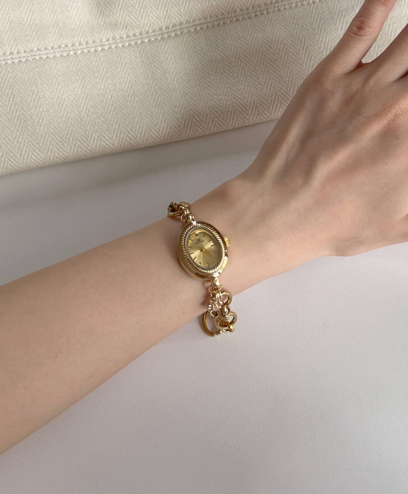 May include: A gold wristwatch with an oval face and a decorative link bracelet. The watch has a gold dial and a detailed bezel. The bracelet includes ornate heart-shaped links and a small charm. The watch is displayed on a wrist.
