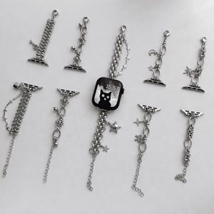 May include: A collection of silver-toned Apple Watch charms and bands. The bands feature various chain designs, charms, and decorative elements, including stars, bells, and a black watch face with a cat graphic.