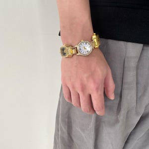 May include: A gold-tone watch with a white face and a square design. The watch has a chain link band and is adorned with small rhinestones.