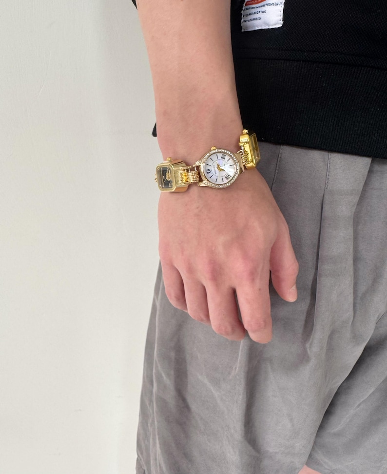 May include: A gold-tone watch with a white face and a square design. The watch has a chain link band and is adorned with small rhinestones.