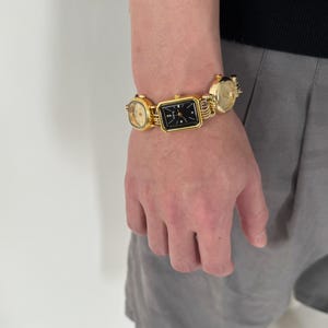 May include: A gold-tone bracelet with three square-shaped watches attached. The watches have black faces and gold-tone hands.