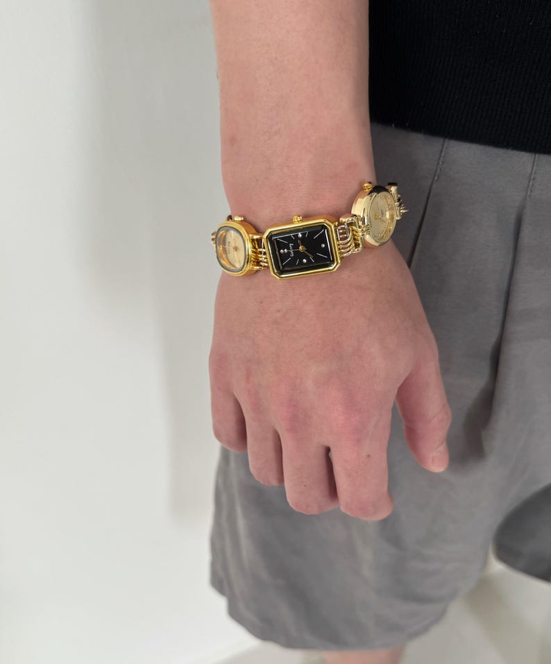 May include: A gold-tone bracelet with three square-shaped watches attached. The watches have black faces and gold-tone hands.